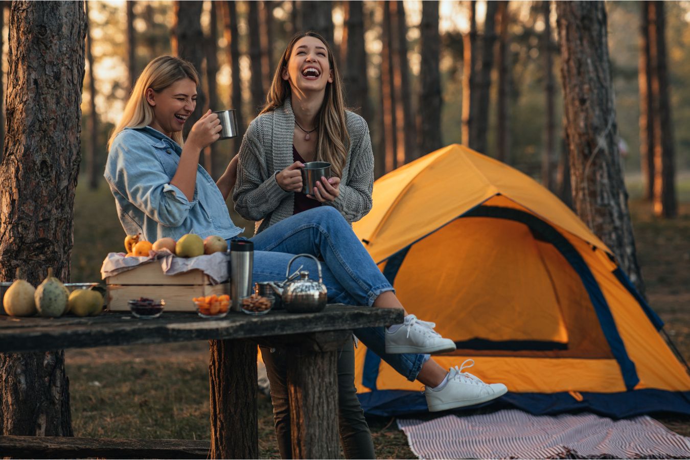 Zwei Frauen lachen vor einem Zelt im Wald. Sie trinken Tee beim naturnahen Camping