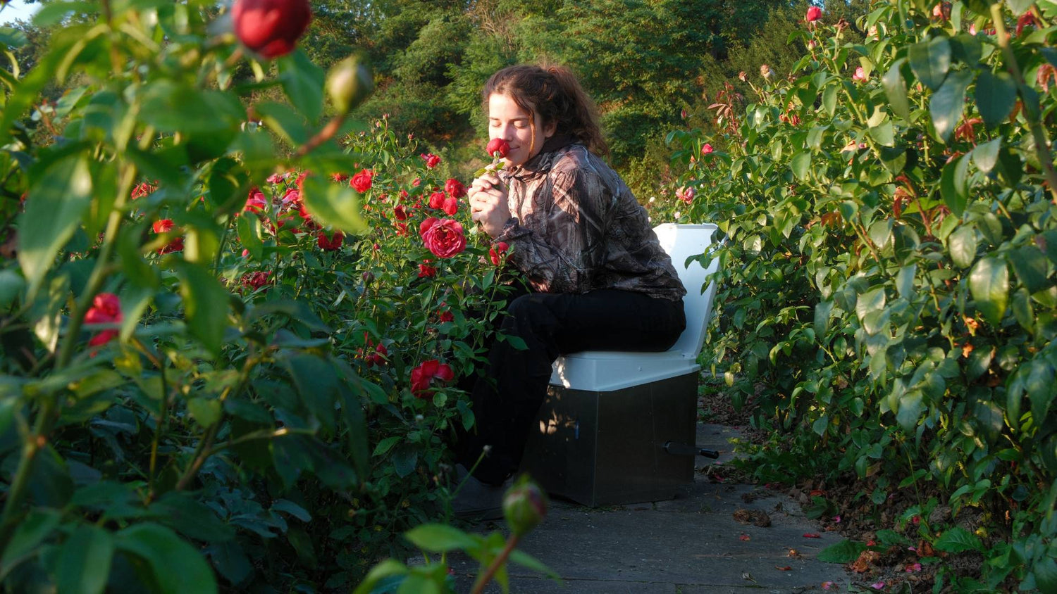 Eine Frau sitzt in der Natur auf einer Trockentoilette für Wohnmobile und riecht an einer Rose.