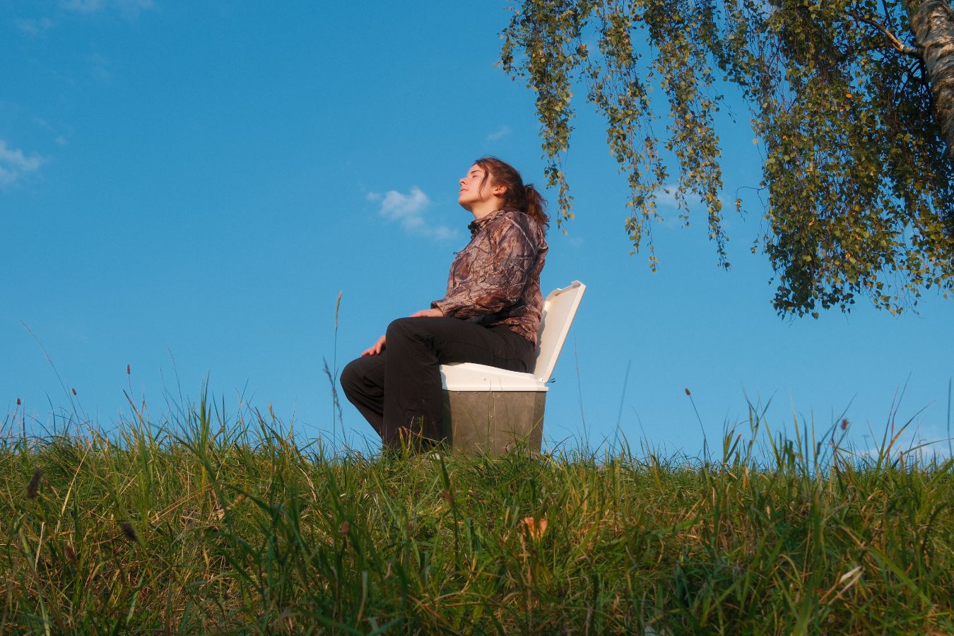 Eine Frau sitzt bei blauem Himmel auf einer Trockentoilette auf einer Wiese. Diese eignet sich für den Einsatz beim Camping ohne Abhängigkeiten.