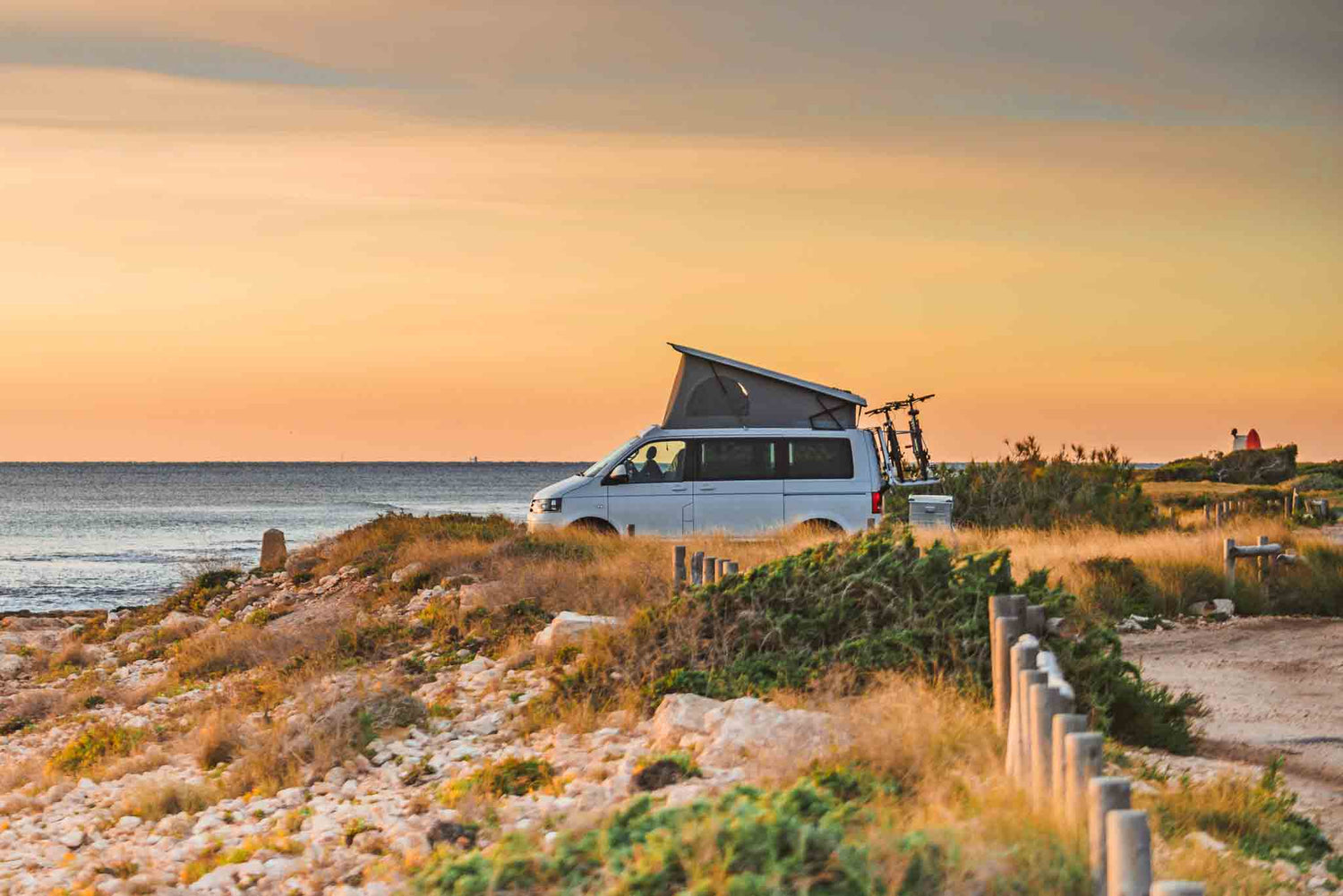 Ein VW California Camper steht im Sonnenuntergang in der Natur an der Küste.